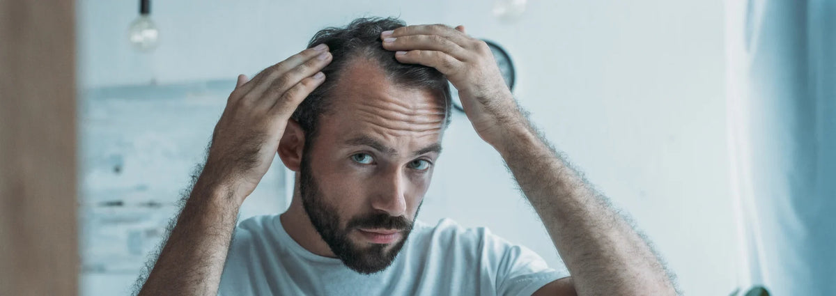 A man with a receding hairline and beard holds his head in frustration, wearing a light gray t-shirt in a brightly lit room.